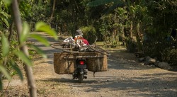 Man on a motorcycle on a rural road on the island of Bali, Indonesia. He is carrying a bicycle and two large baskets full of miscellaneous items. Indonesians carry large loads on their motor scooters
