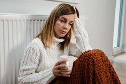 Worried sad woman sits under blanket near heating radiator with cup of tea, Rising costs in private households for gas bill due to inflation and war, Energy crisis