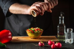 Chef in a restaurant kitchen adds peppers to a fresh vegetable salad. Close-up of a cook hands holding a mill. Cooking healthy and tasty food