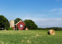 Bright red barn with barn quilt, a silo & round hay bales. Colorful small family farm surrounded by green fields and trees. Concepts of farming, agriculture, Americana, agribusiness, agritourism