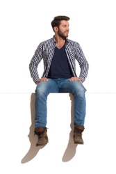 Young man in boots, jeans and unbuttoned lumberjack shirt is sitting on a top, smiling and looking away. Full length studio shot isolated on white.