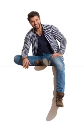 Relaxed young man in boots, jeans and unbuttoned lumberjack shirt is sitting on a top, smiling and looking at camera. Full length studio shot isolated on white.