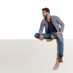 Relaxed young man in boots, jeans and unbuttoned lumberjack shirt sits on a top, smiles and looks away. Full length studio shot isolated on white.
