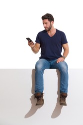 Focused man in boots, jeans and unbuttoned lumberjack shirt is sitting on a top, holding telephone and reading. Full length studio shot isolated on white.