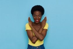 Smiling young black woman with short afro hair is showing letter X with arms crossed. Front view. Waist up studio portrait on blue background.