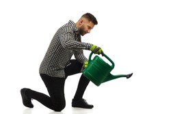 Young focused man in lumberjack shirt is kneeling and watering by using a watering can. Side view. Full length studio shot on white background.