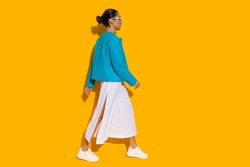 Young woman in blue jacket, white baggy pants and sneakers is walking. Side view. Full length studio shot on yellow background.
