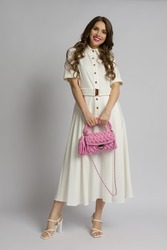 Young woman in white maxi dress and high heels is  holding a pink braided purse. Front view. Full length studio shot.