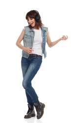 Young woman in jeans vest, black boots and headphones is singing, listening to the music and playing air guitar. Full length studio shot isolated on white.