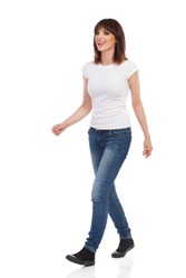 Young woman in jeans and white t-shirt is walking, looking away and talking. Full length studio shot isolated on white.