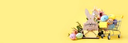 Easter composition with colorful eggs in shopping cart, wooden bunny and spring flowers on yellow background. Banner. Copy space