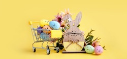 Easter composition with colorful eggs in shopping cart, wooden bunny and spring flowers on yellow background. Banner.