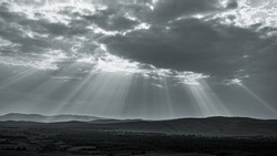 Monochrome photo of bright pillars of sunlight pass through gloomy clouds. Holy Light comes from heaven on Earth. Visible sun light columns above dark mountain landscape in black and white tones
