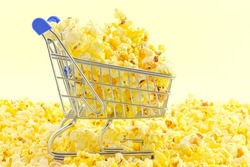 Small shopping cart fully filled with freshly made popcorn among popcorn against yellow background. Unhealthy food concept.