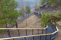 Siberian landscape with winding metal staircase with wooden railing to Torgashinsky ridge in Krasnoyarsk, Russia. Trekking mountain route in springtime day. Mountain slope overgrown with pines