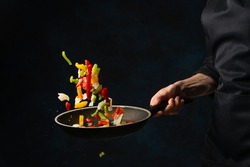 Chef hand throws up frying mix of colored vegetables above the pan on dark blue background. Backstage of cooking meal. Frozen motion. Food banner concept.