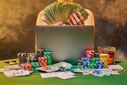 A woman is sitting at the poker table with cash bills in her hands. In front of her is an open laptop. Cards are laid out on the table and multi-colored chips lie in piles. Gambling, online casino.