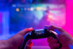 Close-up. Gamepad in the hands of a gamer. Plays video games on the big screen TV. Neon lighting. Video games, communications, cyberspace, eSports, youth culture.