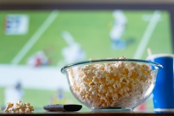 A large bowl of popcorn and a TV remote against the background of a large TV screen with a sports match. Watching sports programs on TV - football, basketball, American football. fast food, snacks.