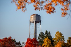 A vintage steel small town water tower standing in morning light and blue skies surrounded by fall color autumn trees
