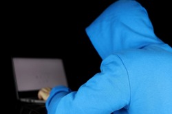 Cybercrime, hacking and technology crime. Man in blue hoodie sitting with laptop on black background, concept of torrent piracy and illegal content