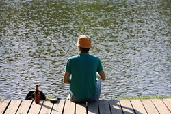 Fisherman sitting on wooden boards with a fishing rod, rear view. Man angling on lake coast at summer