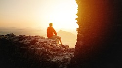 Male traveler sitting on high rock cliff spending time for recreating in nature environment, hipster guy enjoying sunrise during. Discovery, Travel, Adventure

