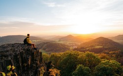 Confident man sitting on the edge. Amazing view  in hilly landscape far from people. Tourist student is relaxing in nature during sunset. Traveling sitting in mountains. Adventure, Art, Travel and Hike concept.