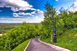 Footpath around the monument in the shade of green trees with lush foliage and a cast-iron black street lamp against a blue sky with white clouds. Park of Salavat Yulayev, Ufa, Bashkortostan, Russia.