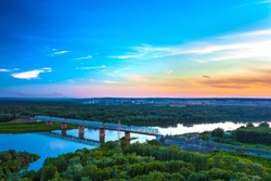 Railway bridge is reflected over the calm White River, surrounded by a green lush forest on the outskirts of the city under the evening blue sky at dusk. Sunset over Ufa, Bashkortostan, Russia.
