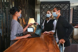 Couple and receptionist at counter in hotel wearing medical masks as precaution against virus. Young couple on a business trip doing check-in at the hotel