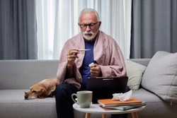 Sick elderly man checking his temperature suffering from seasonal flu or cold and using digital tablet for entertainment or online doctor consultation. Ill senior feel unhealthy with influenza at home