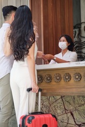 Business partners or couple and receptionist at counter in hotel wearing medical masks as precaution against virus. Young asian couple doing check-in at the hotel and receiving room key