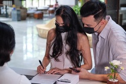 Business partners or couple and receptionist at counter in hotel wearing medical masks as precaution against virus. Young asian couple filling up information form at reception counter in hotel