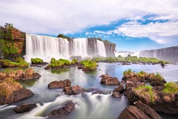 Iguazu waterfalls in Argentina. Panoramic view of many majestic powerful water cascades with mist and clouds. Panoramic image of Iguazu valley with grass and stones in calm water.