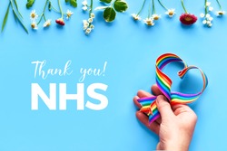 Thank you NHS, doctors, nurses, medical teams and key workers in England and UK! Hand holding rainbow ribbon in heart shape on blue mint background with flowers, trendy flat lay, text 