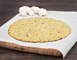 Plain cauliflower pizza crust on a piece of parchment paper on a cutting board.  Selective focus on front edge of crust.
