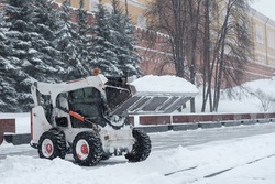 A small loader excavator bobcat removes snow from the sidewalk near the Kremlin walls during a heavy snowfall. Snowflakes are flying in the air.