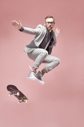 Young handsome serious man with glasses, brown hair and beard, wearing light grey suit and sneakers, jumping with the skateboard and flying in the air on light pink background 