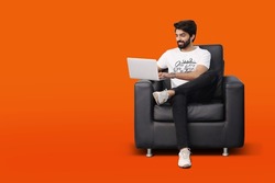 Happy young bearded man sitting on black sofa couch chair using laptop on orange background - Pakistani Indian South Asian