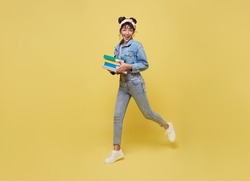 Happy Asian girl holding book and jumping over yellow background. back to school concept.