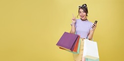 Cheerful happy teen asian woman enjoying shopping, she is carrying shopping bags and hand pointing isolated over yellow background.