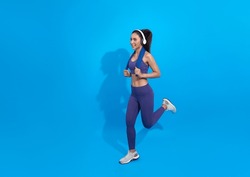 young asian fitness woman in sportwear running wearing headphones listening to music isolated on blue copy space studio background . excited runner
