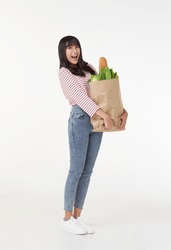 Full body smiling happy young asian housewife hold paper bag with fresh food products isolated on wite background studio. shopping in supermarket