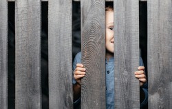 Happy smiling white boy looks out of the crack of a wooden fence. Childish curiosity. Espionage. Rural life. Child development, curiosity. The boy peered through the crack.