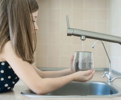 Little girl open a water tap with her hand holding a transparent glass. Kitchen faucet. Filling cup beverage. Pouring fresh drink. Hydration. Healthcare. Healthy lifestyle. World water day