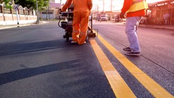 Rear view of 2 road workers with thermoplastic spray marking machine working to paint yellow lines on asphalt road surface with flare light in evening time, selective focus 