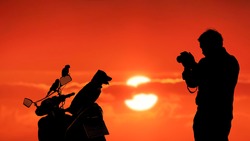Silhouette young man using camera to taking picture of his pets [puppy and 2 parrots] on motorcycle with blurred colorful sunset sky background