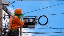 Technician on wooden ladder Checking Fiber Optic Cables in Internet Splitter Box on Electric Pole against blue sky background