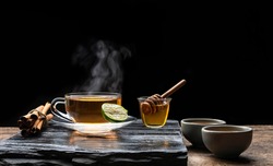 Aromatic herbal hot tea in glass teacup with steam and honey on black stone plate in dark background